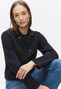 Donkerblauw gebreid dubbelgeknoopt jasje met ronde kraag, zwarte knopen en lange mouwen. Draagt blauw spijkerbroek en toont een casual pasvorm.