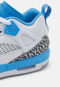 Zapatilla deportiva blanca con acentos azules, mediasuela gris texturizada, detalles de malla y marca prominente en la lengüeta. Presenta superposiciones de plástico.