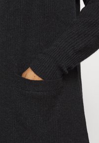 Černý pletený svetr zblízka, s riblovanou texturou, dlouhými rukávy a postranní kapsou, ukazující měkkou tkaninu a čistý design.