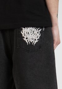 Un pantalon en denim noir présente un graphique brodé blanc sur la poche arrière. Le tissu semble légèrement décoloré, ajoutant une texture.
