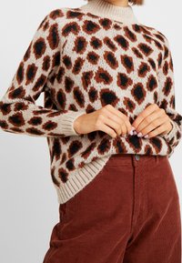 Strickpullover mit beigem Grundton und rostfarbenen sowie schwarzen Leopardenflecken, gerippter Hals- und Saumabschluss, kombiniert mit rostfarbener Cordhose.