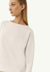 Light beige oversized sweatshirt with a wide neckline, ribbed cuffs, and a subtle brand logo on the hem. Soft cotton blend texture.