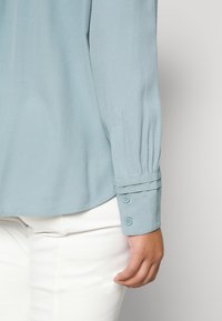 Blouse bleu clair avec une texture lisse, dotée de manches longues, de poignets boutonnés et de douces plis aux épaules. Associée à un pantalon blanc.