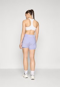 Bermudas deportivas de color púrpura claro con cintura elástica, acompañadas de un sujetador deportivo blanco de espalda nadadora. Tejido suave, corte casual, sin patrones visibles.