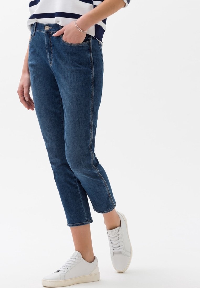 Personne portant un jean bleu, des baskets blanches et une chemise blanche à rayures bleu marine, debout avec une main dans la poche devant un fond uni.