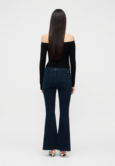 Pikkade sirgete juustega naine, kes kannab musta õlgadeta pluusi, tumelgeid lehvivaid teksapükse ja valgeid kontsakingi, seisab seljaga vaataja poole.