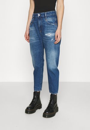 Personne portant un jean bleu délavé et ajusté, des bottes à plateforme noires à lacets, et un haut noir sans manches, debout devant un fond uni.