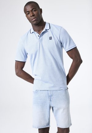 Man draagt een lichtblauw gestructureerd poloshirt en lichte denimshorts, staand met handen achter de rug tegen een witte achtergrond.