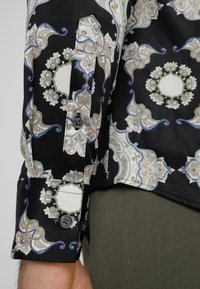 Chemise noire à motifs avec des dessins floraux et géométriques complexes en beige, bleu et vert. Comprend des boutons noirs sur les poignets.