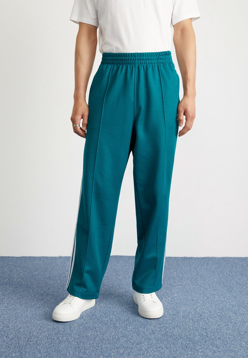 Teal treningsbukser med elastisk midje, med hvite sidelister og løs passform. Brukt med hvite joggesko på et blått gulv.