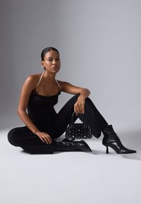 Svart bodysuit med rynket design, parret med vide bukser. Holder en liten svart veske med sølvnitter, iført høye svarte støvler.