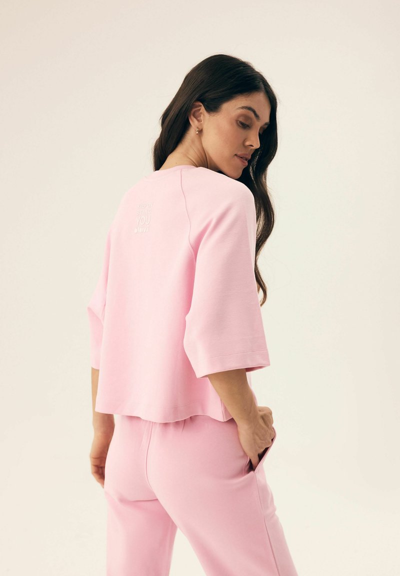 Jasnoróżowy bawełniany sweatshirt z rękawami 3/4 i skróconym krojem. Wyróżnia się wytłoczonym napisem „Być Sobą” z tyłu. Połączony z dopasowanymi spodniami.