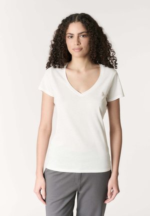 Giovane donna con capelli ricci che indossa una maglietta bianca a scollo a V e maniche corte e pantaloni grigi, in piedi davanti a uno sfondo semplice.