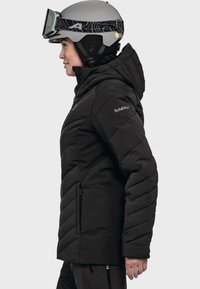 Schwarze isolierte Jacke mit einem Chevron-Design, seitlichen Taschen und einer Kapuze. Trägt einen grauen Skihelm und Skibrille, zeigt ein aerodynamisches Profil.