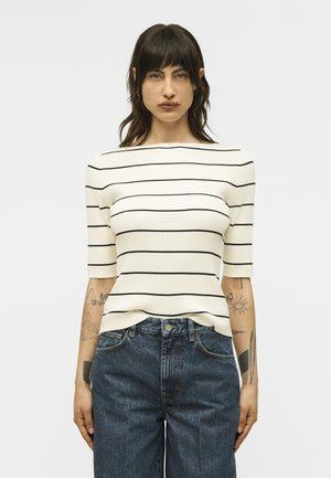 Γυναίκα με τατουάζ που φοράει λευκό κοντομάνικο τοπ με μαύρες οριζόντιες ρίγες και μπλε ψηλόμεσα τζιν, στέκεται μπροστά σε απλό φόντο.