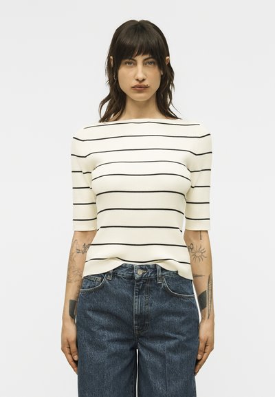 Γυναίκα με τατουάζ που φοράει λευκό κοντομάνικο τοπ με μαύρες οριζόντιες ρίγες και μπλε ψηλόμεσα τζιν, στέκεται μπροστά σε απλό φόντο.