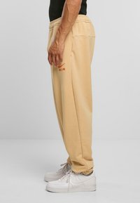 Beige sporta bikses ar brīvu piegriezumu, elastīgu jostu, sānu kabatām un sarkanu izšūtu 'AIR' logo kreisajā augšstilbā.