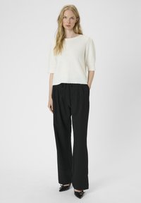 Vrouw die een witte korte mouw trui, losse zwarte broek en zwarte puntige schoenen draagt, staande tegen een effen achtergrond.