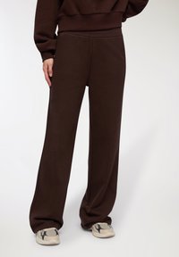 Pantaloni della tuta marroni realizzati in un tessuto morbido, caratterizzati da una vestibilità comoda, un elastico in vita e gambe larghe. Abbinati a sneakers beige.