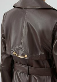 Bruine leren trenchcoat met een grote kraag, een ceintuur in de taille en een gouden kettingaccent op de rugzijde. Soepele textuur, aangepast pasvorm.