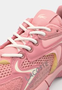 Růžová sportovní bota s panelu z síťoviny a semiše, bílé šňůrky a výrazným logem Lacoste. Má zaoblený tvar a strukturované detaily.