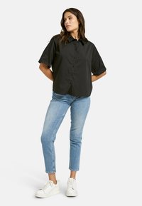 Black short-sleeve button-up shirt with a collar, paired with light blue slim-fit jeans and white sneakers, standing against a plain background.