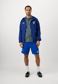 Mėlynas sportinis švarkas su auksiniais akcentais, dryžuota marškiniai, mėlyni šortai, balti kojinės ir juodi batai, su Italijos logotipų detalėmis.