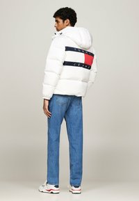 Giacca a vento bianca con cappuccio, caratterizzata da una fascia logo a strisce blu e rosse sulla schiena, abbinata a jeans blu e sneakers bianche.