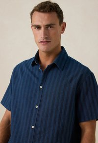Korte mouwen, knoopshirt in marineblauw met verticale strepen. Gemaakt van lichte stof en heeft een klassieke kraag en plastic knopen.