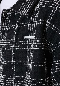 Zwarte textuurjack met een wit geruit patroon en een klein metalen label met de tekst "PROJECT X PARIS" op de borst.