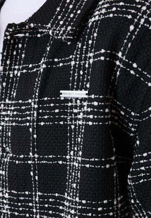 Zwarte textuurjack met een wit geruit patroon en een klein metalen label met de tekst "PROJECT X PARIS" op de borst.