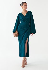 Vestido envolvente de manga larga en color azul verdoso con un escote en V profundo y una abertura lateral. Presenta una tela suave y detalle fruncido en la cintura. Tacones con tiras doradas.