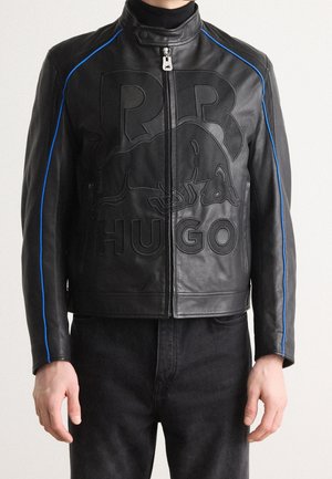 Musta nahkatakki, jossa kohokuvioitu Red Bull -logo, siniset somisteet hihoissa, vetoketju edessä, päällä musta pooloneule ja mustat housut.