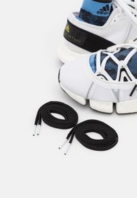 Baskets de sport blanches avec une tige en mesh, doublure de chaussette bleue, accents noirs et semelle blanche épaisse. Comprend deux lacets noirs avec des embouts blancs.