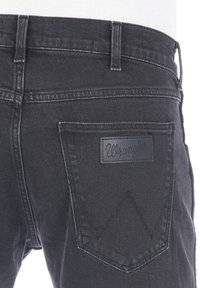 Jeans in denim nero con una tasca posteriore caratterizzata da una toppa con il logo Wrangler in pelle nera e un dettaglio di cuciture distintive sulla tasca.