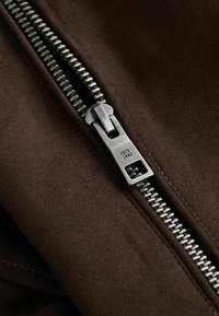 Brown suede fabric with a silver metal zipper. The zipper is partially opened, showcasing teeth and a rectangular pull tab.