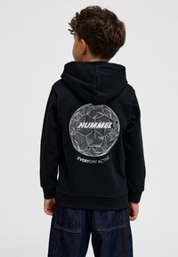 Zwarte hoodie met een ronde witte graphic op de achterkant, voorzien van lijnen en de tekst "HUMMEL EVERYDAY ACTIVE." Zacht materiaal en een kangoeroezak.