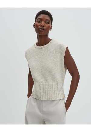 Frau trägt ärmellosen beigen Strickpullover und hellgraue Hose, steht mit Händen in den Taschen vor einfarbigem Hintergrund.