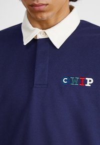 Mörkblå polo-shirt med krämfärgad krage och tre knappslutningar. Har en flerfärgad broderad "CWIP"-logotyp på bröstet.