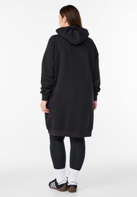 Sort quiltet hættetrøjekjole med rummelig hætte, lange ærmer, ribstrikkede manchetter og en længde lige over knæet. Bær med sorte leggings og sokker.