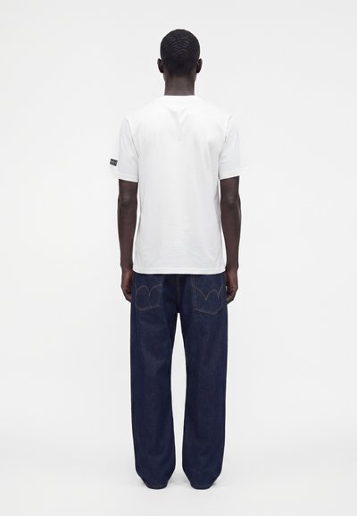 Homme debout de dos, portant un t-shirt blanc uni à manches courtes et un jean ample en denim bleu foncé sur fond blanc.