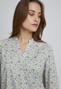 Blumenbluse auf einem hellcremefarbenen Hintergrund mit blauen und rosa Blumen, einem V-Ausschnitt und langen, locker sitzenden Ärmeln.