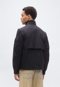 Άνδρας με κοντά σγουρά μαλλιά φοράει μαύρο μπουφάν με αεραγωγό στην πλάτη και μπεζ παντελόνι, στέκεται μπροστά σε απλό ανοιχτό γκρι φόντο.