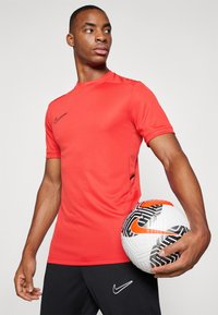 Rød Nike kortermet treningsskjorte med svart logo, strukturert stoff og sidespalter, sammen med en mønstret hvit og svart fotball.