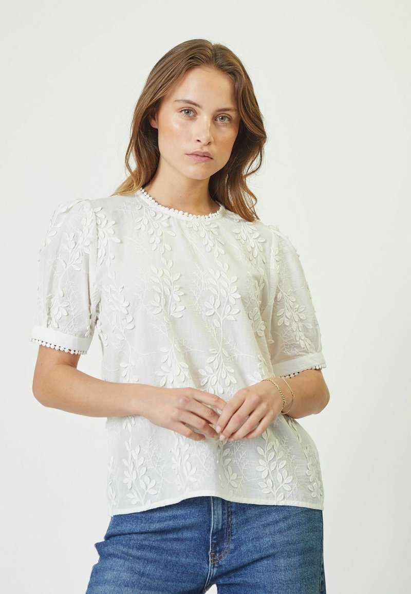 Blusa bianca ricamata con motivi a foglia, maniche a palloncino corte e scollatura a onde, abbinata a jeans di denim blu.