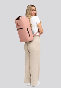 Mochila impermeable rosa con cremalleras negras y una correa ajustable, que presenta una parte superior redonda y un logo minimalista en la parte frontal.