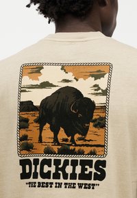 Μπεζ κοντομάνικη μπλούζα με γραφικό ταύρου, περιτριγυρισμένο από περίγραμμα σχοινιού, σε γήινες αποχρώσεις καφέ, πράσινου και πορτοκαλί. Το όνομα της μάρκας "Dickies" από κάτω.