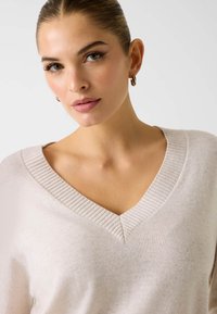 Jasnobeżowy sweter z prążkowanym dekoltem w serek, o miękkiej fakturze i luźnym kroju, noszony przez modelkę z minimalnym makijażem.