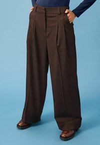 Pruunide triibuliste pükste krookedega laiad jalad, esi-plekkidega, kõrge vöökoht ja küljetaskud, koos tumedate kingadega.