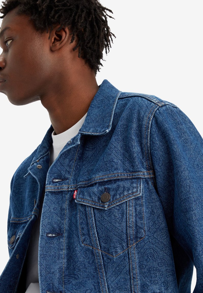 Veste en denim bleu au design classique, avec deux poches poitrine, des boutons en bronze et une texture subtile avec des détails de couture visibles.